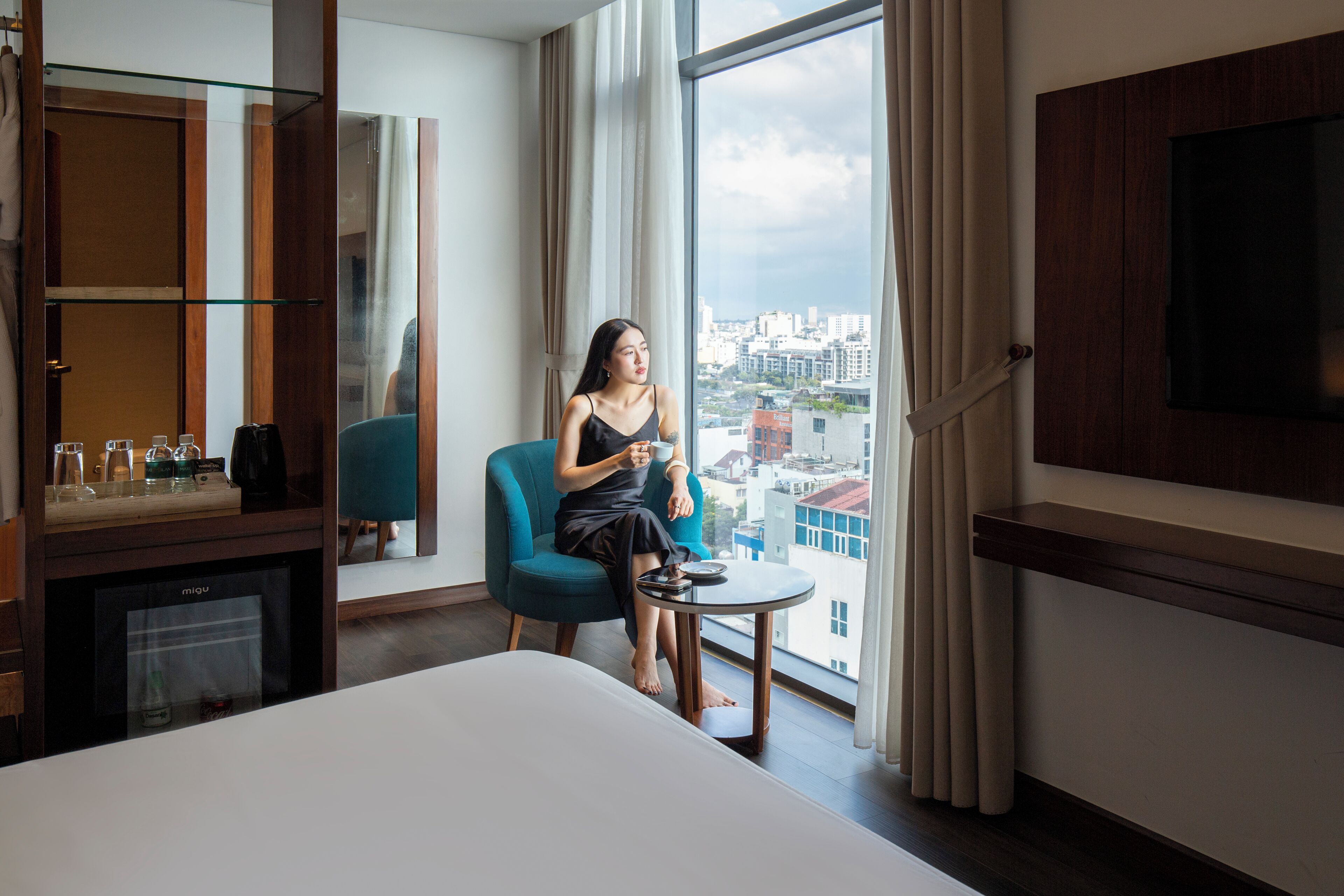 A hotel room with a large window, a bed, a small round table, and a television mounted on the wall.