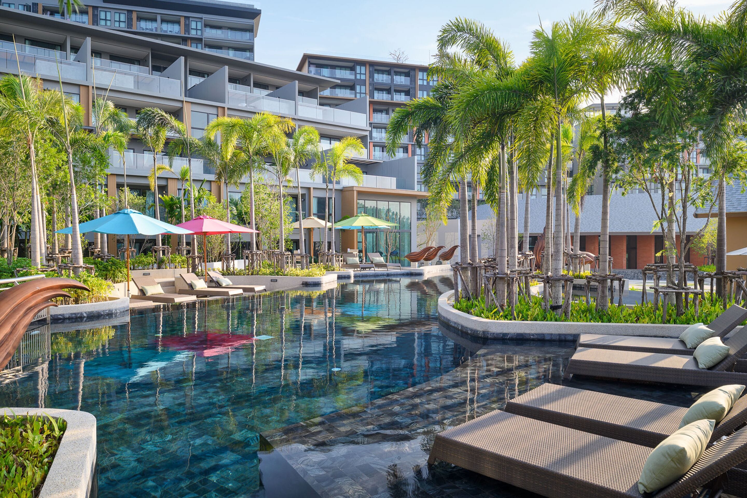 A pool area with lounge chairs and umbrellas, surrounded by palm trees and a modern building.