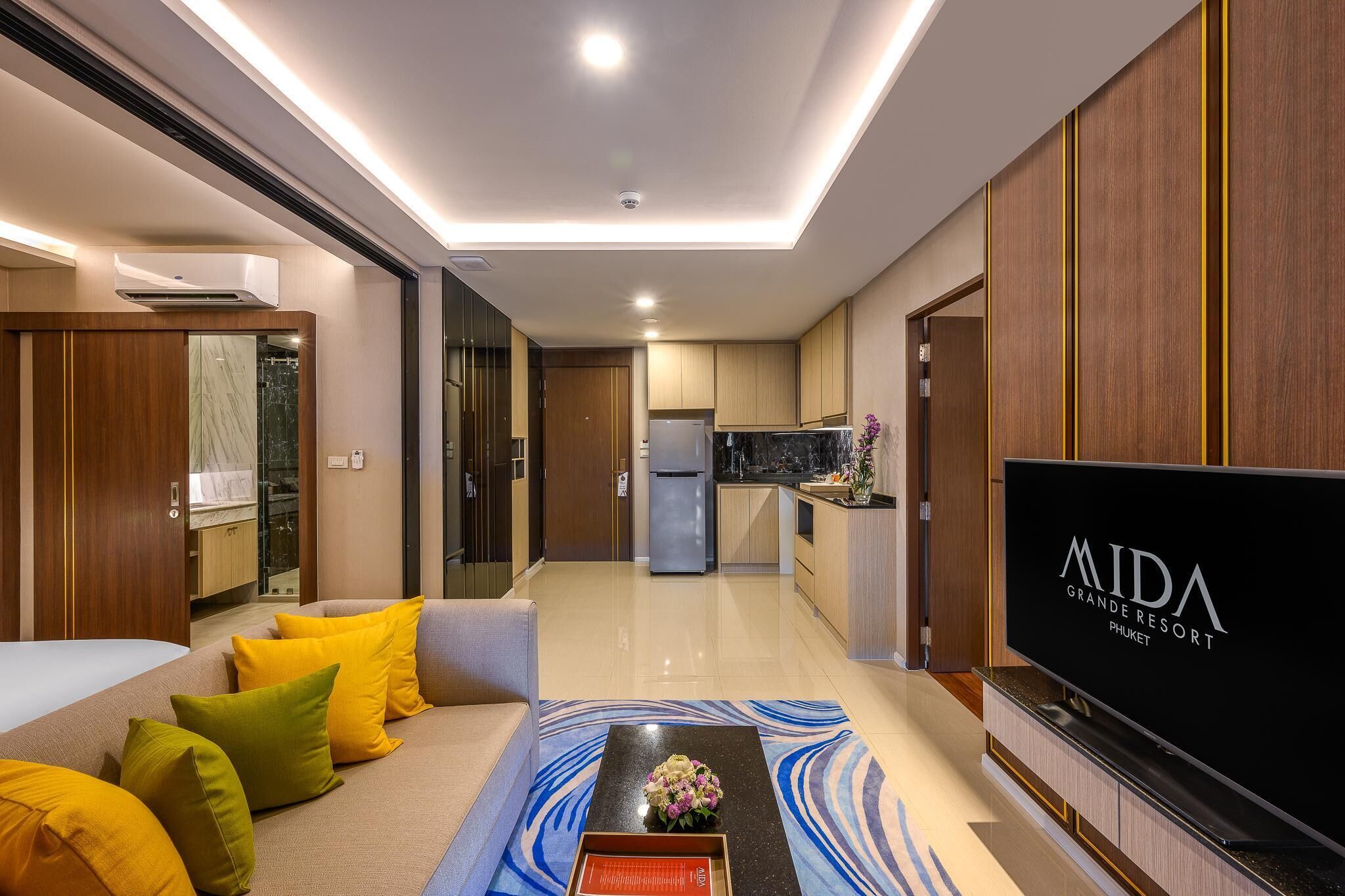 A modern hotel room with a sofa, a coffee table with a vase, a flat-screen TV, and a kitchen area in the background.