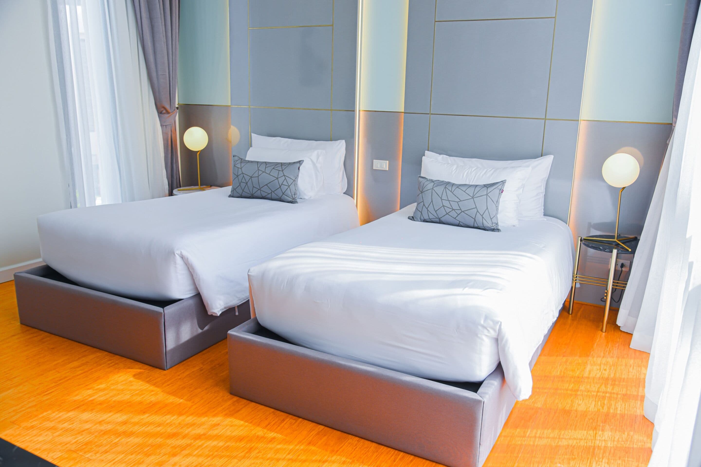 Ein Hotelzimmer mit zwei Betten, jeweils mit grauem Lattenrost und weißer Bettwäsche. Es gibt Nachttische mit Lampen, und der Raum hat ein modernes Design mit einer geometrischen Wandverkleidung.