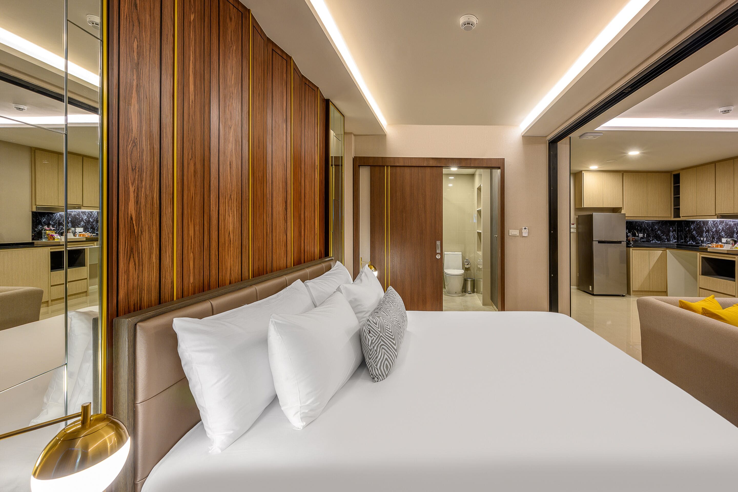 Habitación de hotel moderna con una cama grande, paneles de madera, una cocina al fondo y un baño visible a través de una puerta abierta.