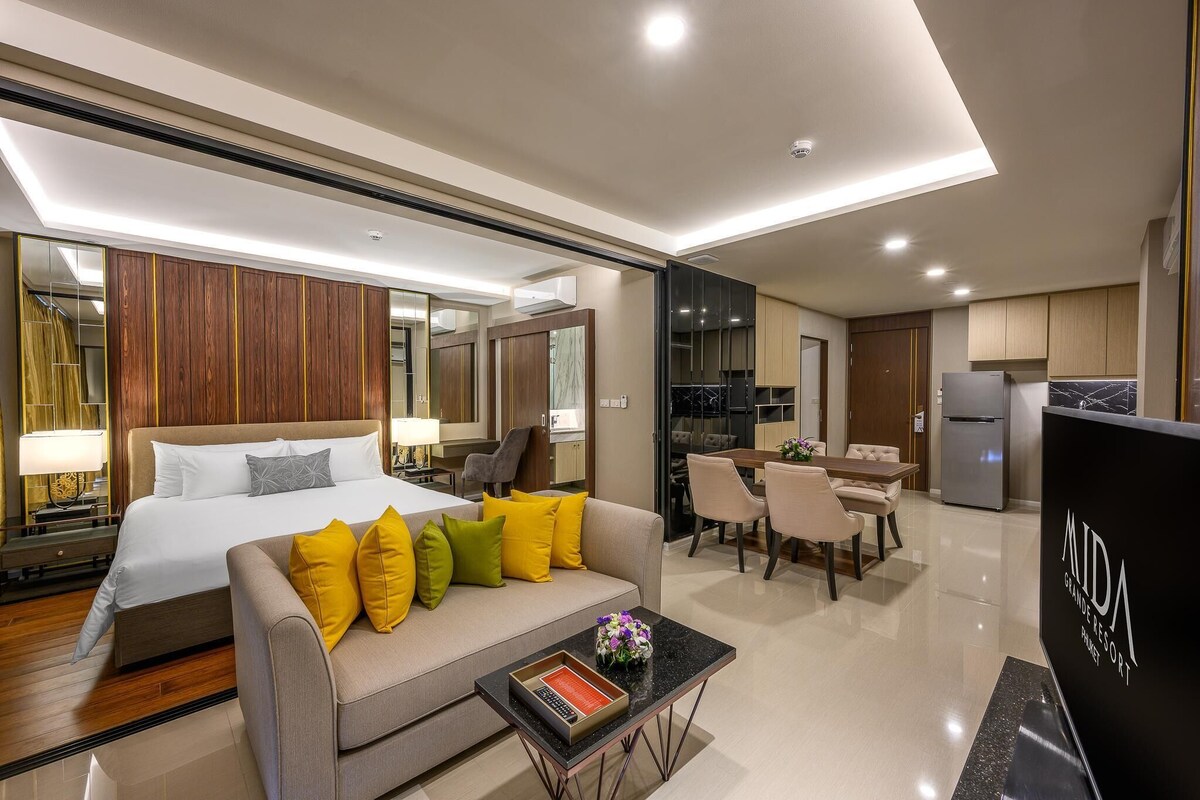 A modern hotel room with a large bed, a sofa with colorful cushions, a dining area, and a kitchen.