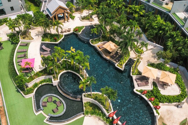 An aerial view of a resort with a central swimming pool, surrounded by palm trees and lounge areas.