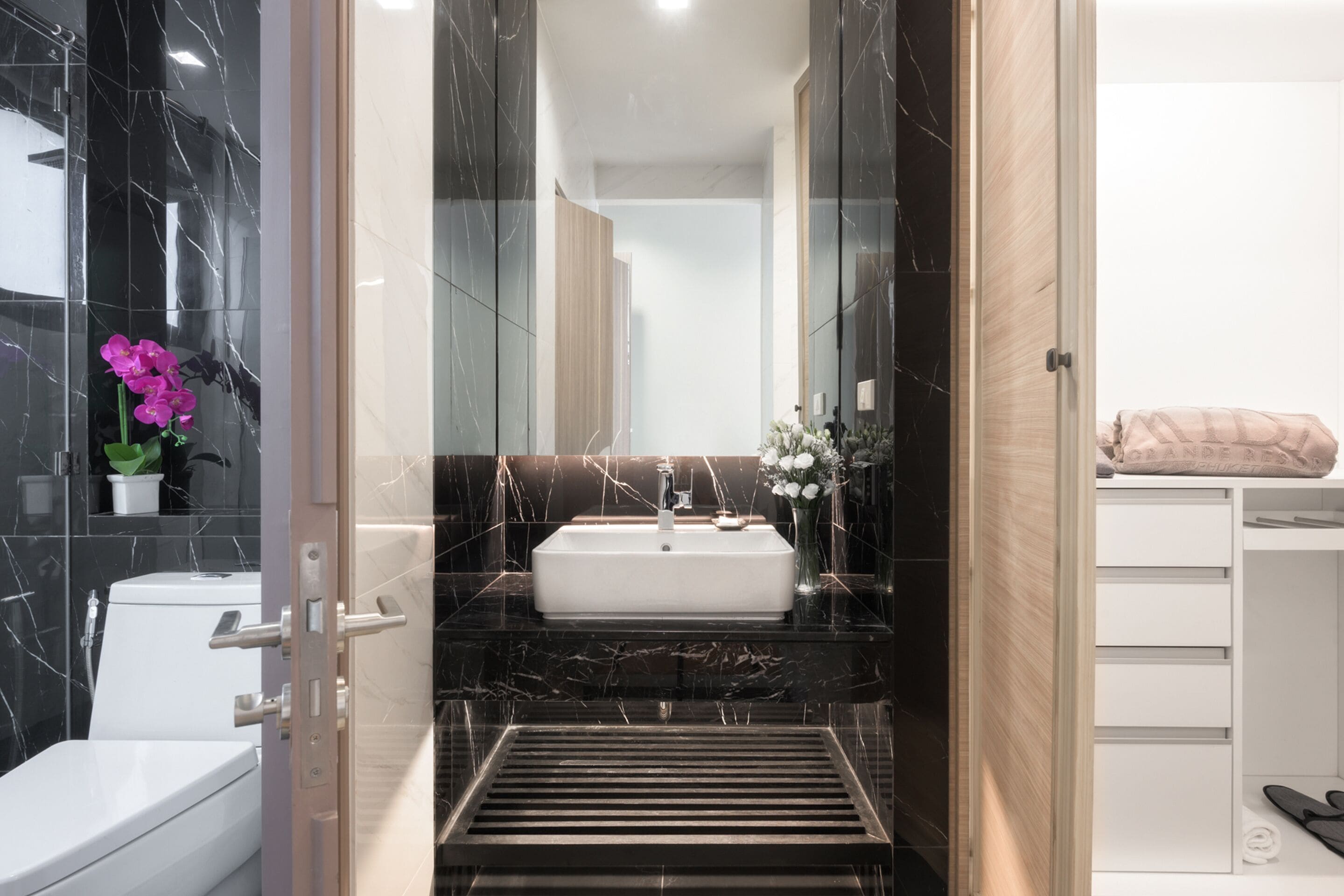 A modern bathroom with a white sink, black marble walls, and a wooden cabinet.