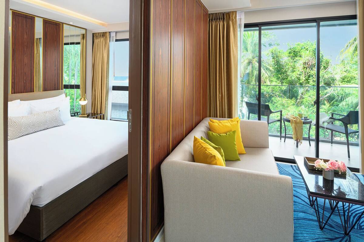A hotel room with a bed, a sofa, a balcony with chairs, and a view of greenery.