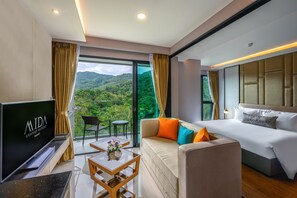 A modern hotel room with a large bed, a sofa, a TV, and a balcony with a view of the mountains. - MIDA Grande Resort Phuket (Choeng Thale)
