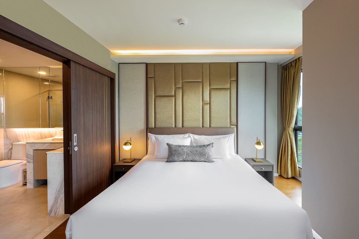 A modern hotel room with a large bed, a bedside table with a lamp, a wardrobe, and a view of the outdoors.