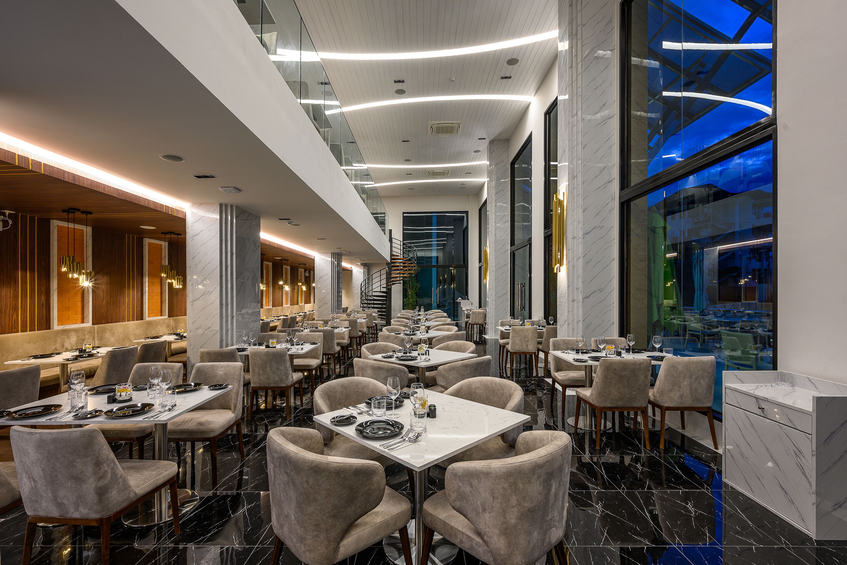 Un restaurante moderno con varias mesas y sillas, piso de mármol y vistas panorámicas de la ciudad a través de amplios ventanales.