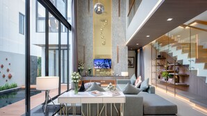 A modern living room with a sofa, a coffee table, and a TV. There is a staircase and a bookshelf in the background. - MIDA Grande Resort Phuket (Choeng Thale)