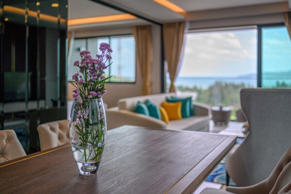A modern living room with a wooden table, a vase with purple flowers, and a view of the outdoors through large windows. - MIDA Grande Resort Phuket (Choeng Thale)