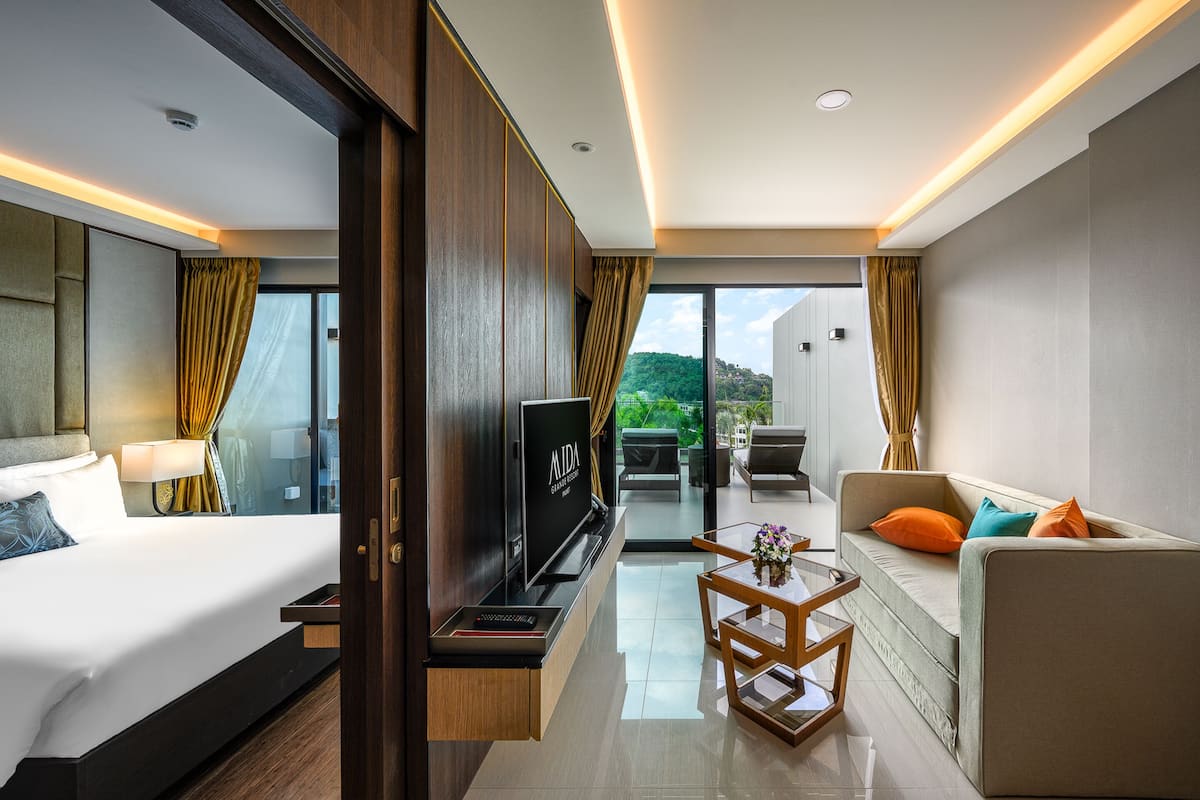 A modern hotel room with a bed, a TV, a sofa, a small table, and a balcony with a view of greenery.