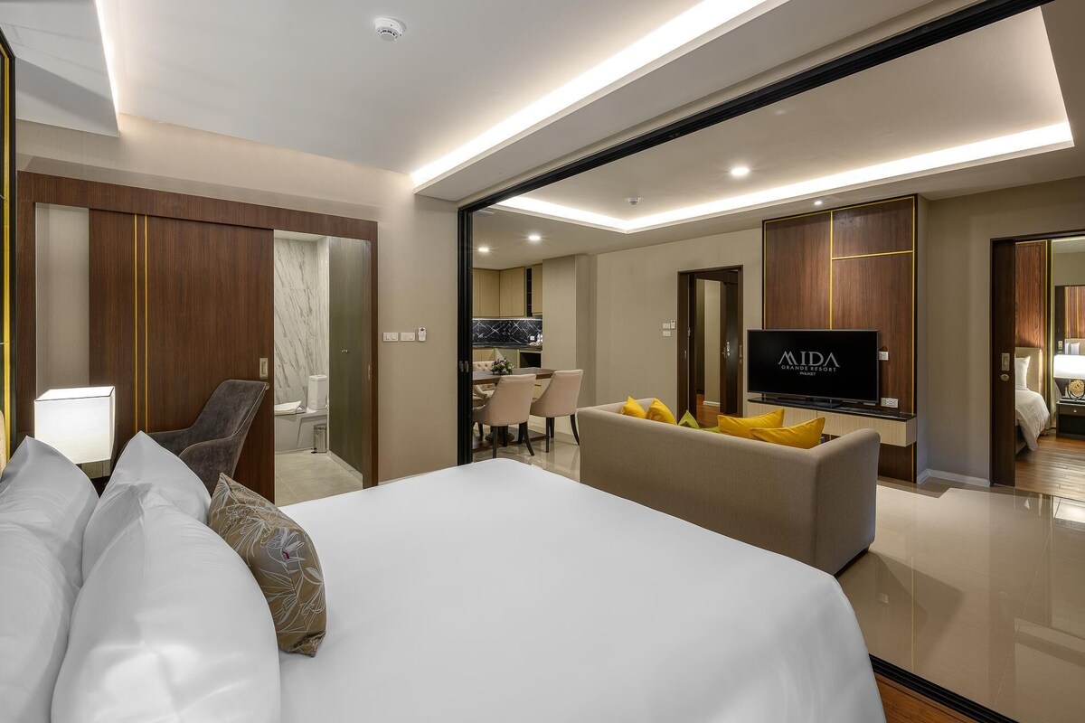 A modern hotel room with a large bed, a sofa, a TV, and a dining area visible through an open door.