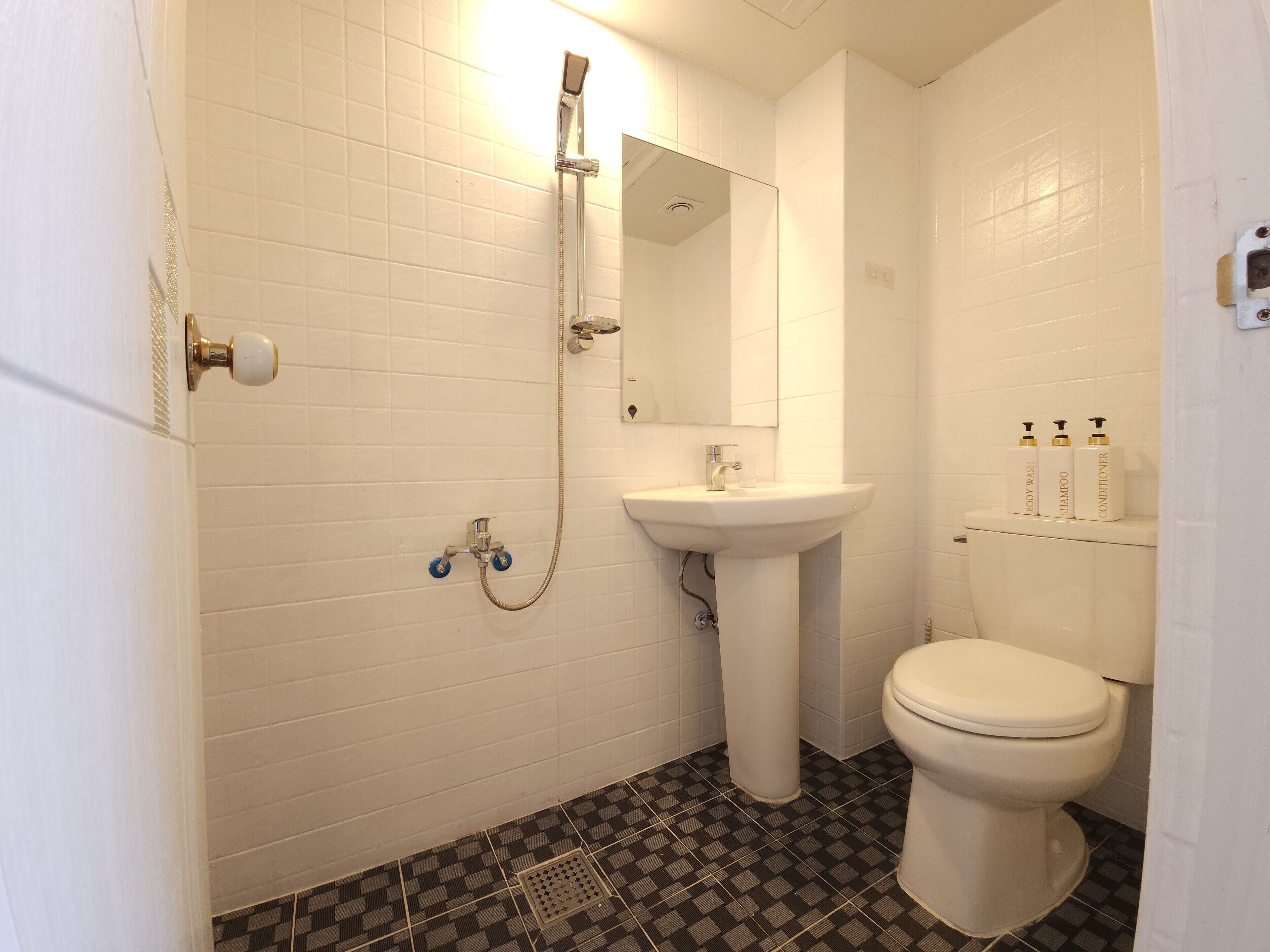 Casa de banho com paredes revestidas a azulejos brancos, uma sanita, uma pia branca e um chuveiro com cabeça de duche manual. Há três frascos de artigos de higiene pessoal na parede.