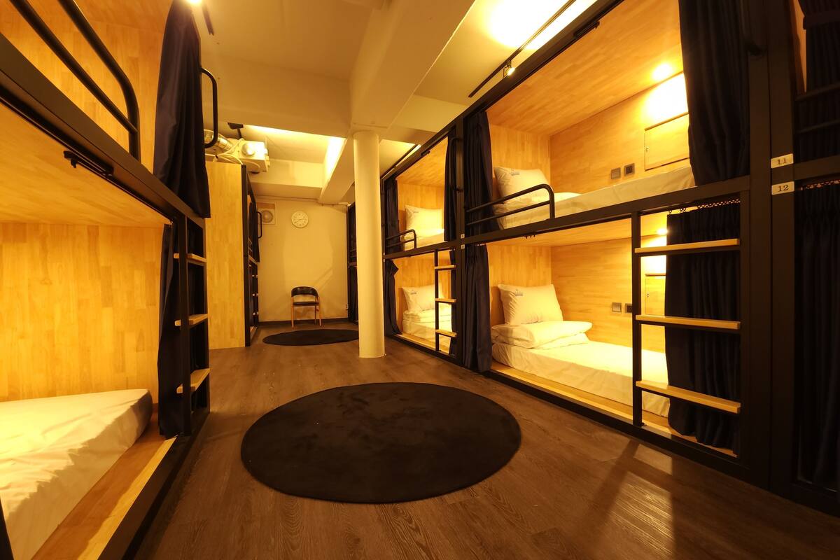 Habitación compacta y bien iluminada con literas, una silla pequeña y una alfombra redonda.