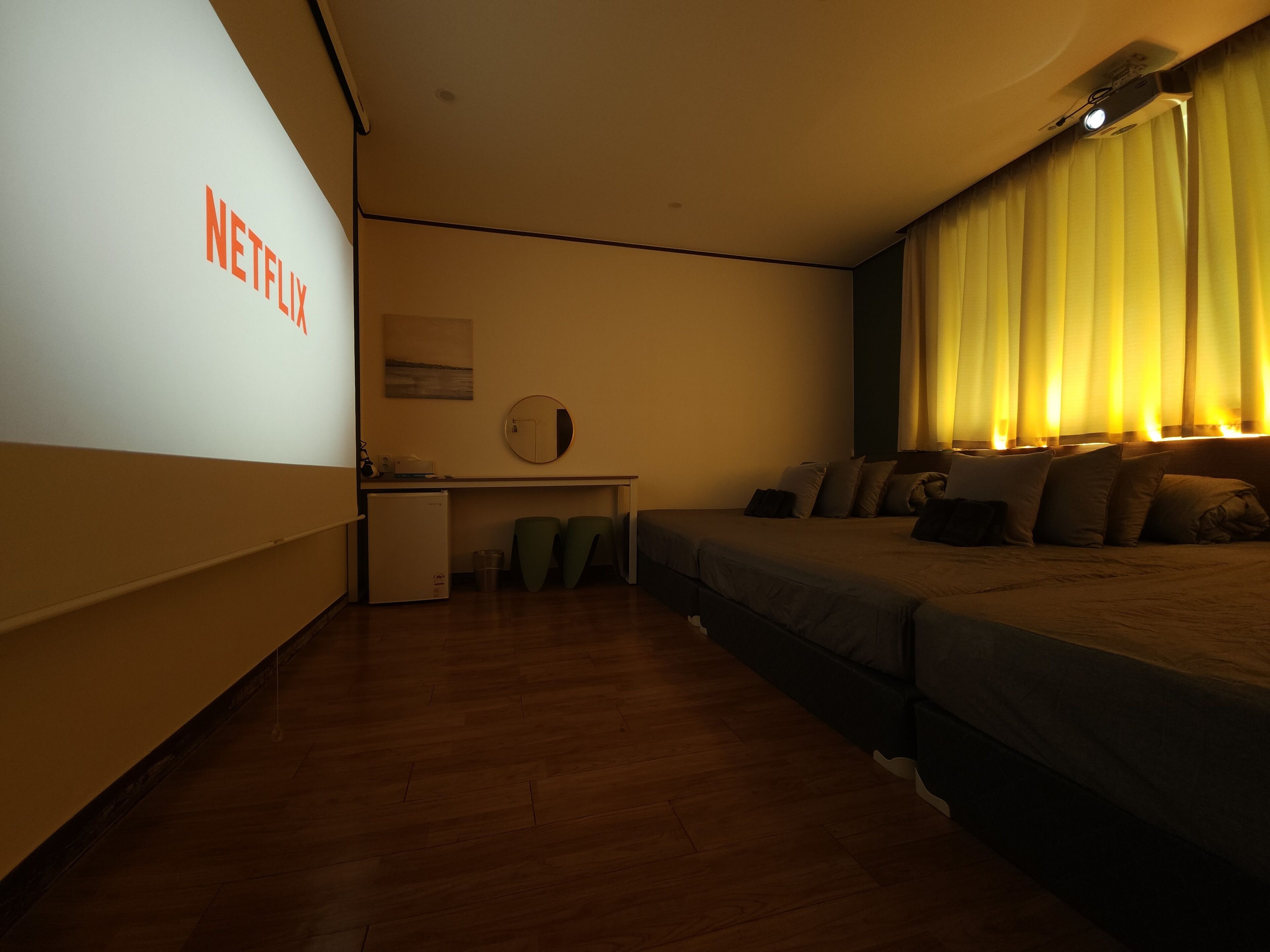 A hotel room with a bed, a desk, a small refrigerator, and a wall-mounted projector displaying the Netflix logo.