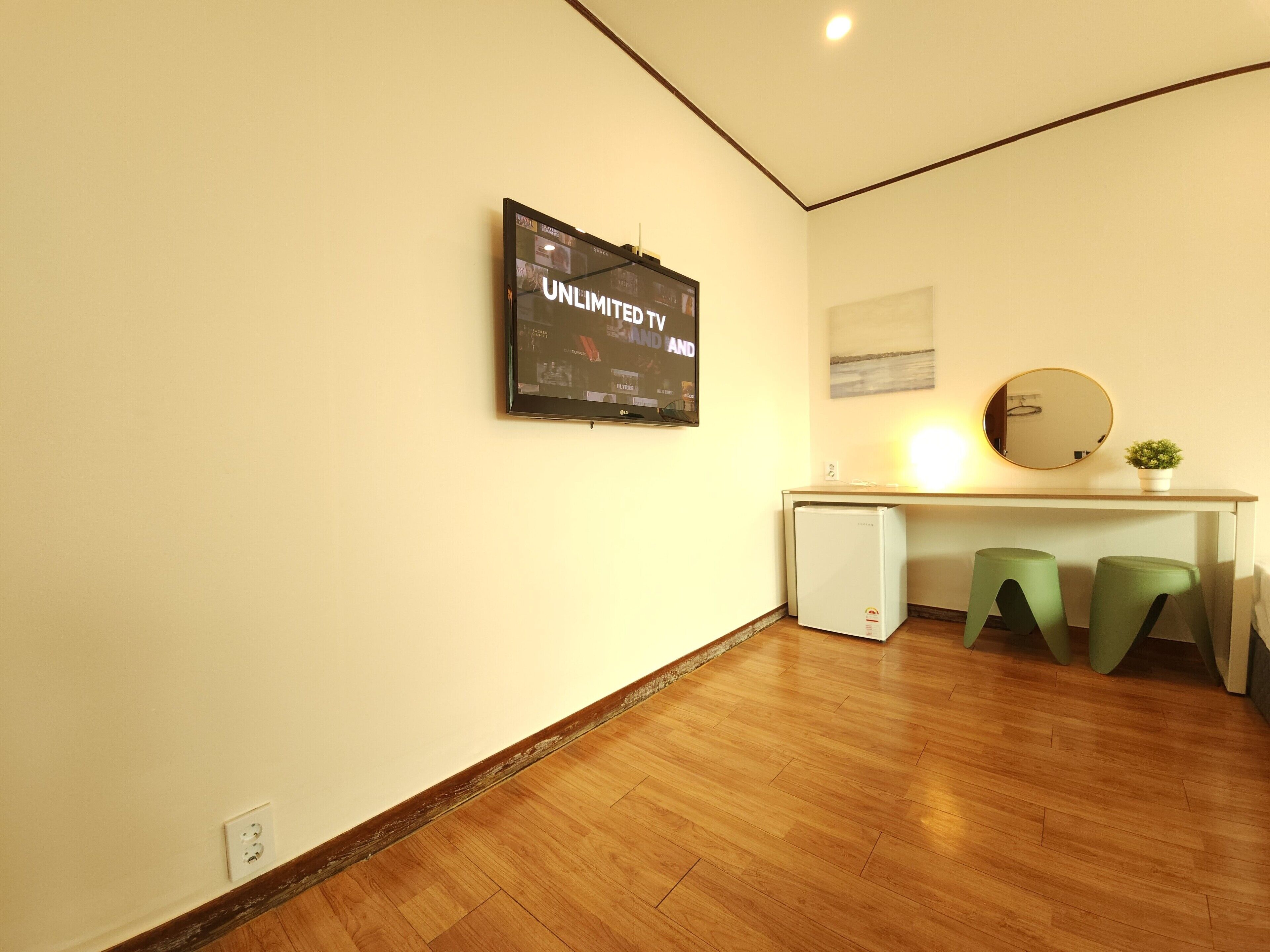 A small room with a wooden floor, a white desk with a computer, two green stools, a small fridge, a wall-mounted TV displaying 'UNLIMITED AND', and a round mirror above the desk.