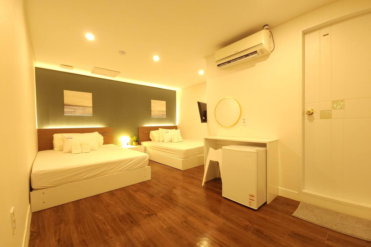 Habitación de hotel con dos camas, un escritorio, un refrigerador pequeño, aire acondicionado y una puerta con felpudo.