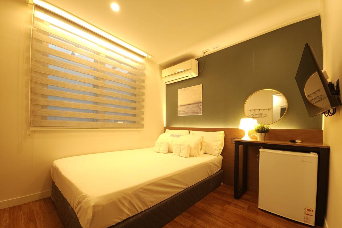 Un dormitorio compacto con cama, un refrigerador pequeño, una mesita de noche con lámpara y un espejo montado en la pared.