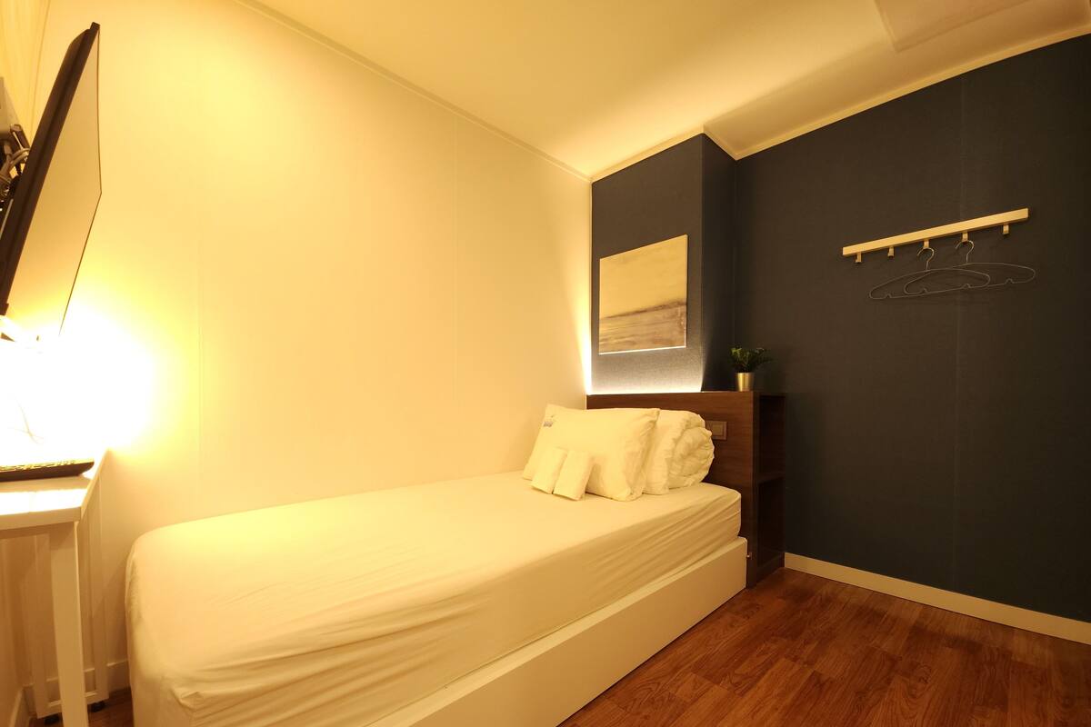 Un dormitorio con una cama individual, un escritorio con lámpara, una pared oscura con un cuadro, y piso de madera.