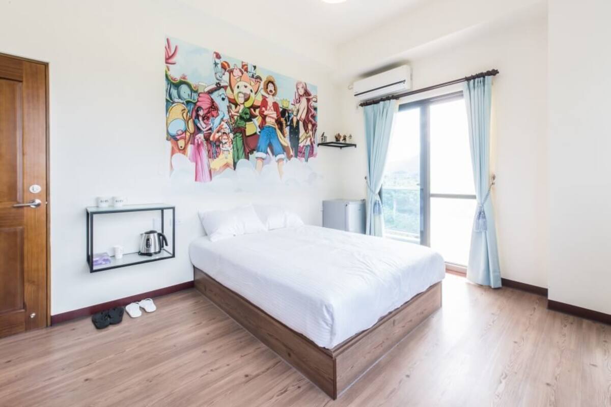 Une chambre avec un grand lit, une étagère fixée au mur avec une bouilloire, une paire de chaussures sur le sol et une fenêtre avec des rideaux.
