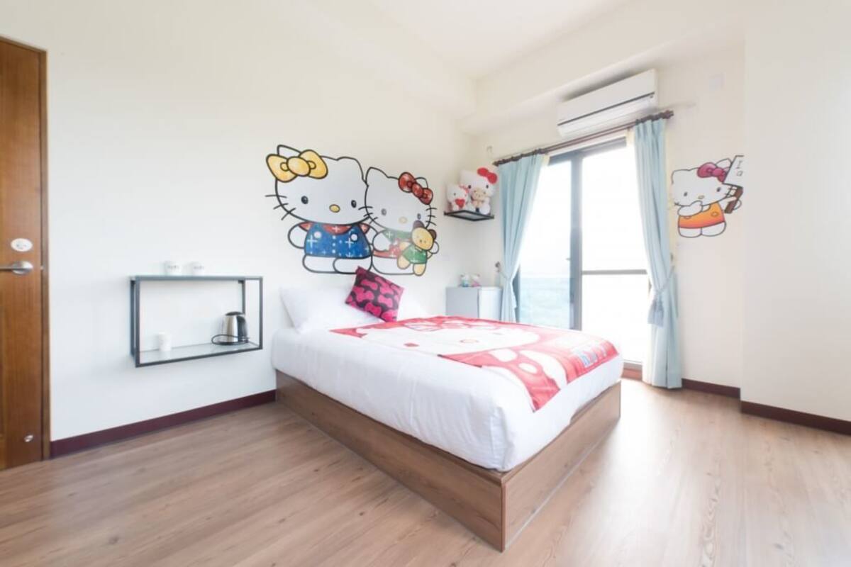 Une chambre avec un lit en bois, des autocollants Hello Kitty sur les murs, une table de chevet et une fenêtre avec des rideaux.