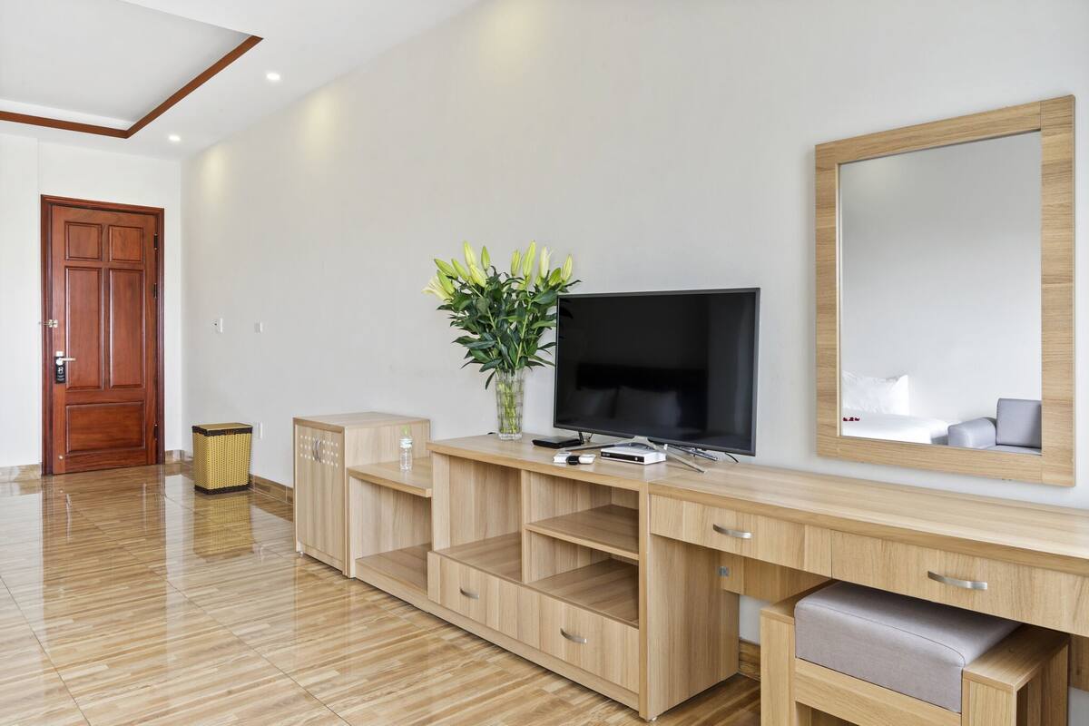 A modern hotel room with a wooden desk, a flat-screen TV, a large mirror, and a potted plant.