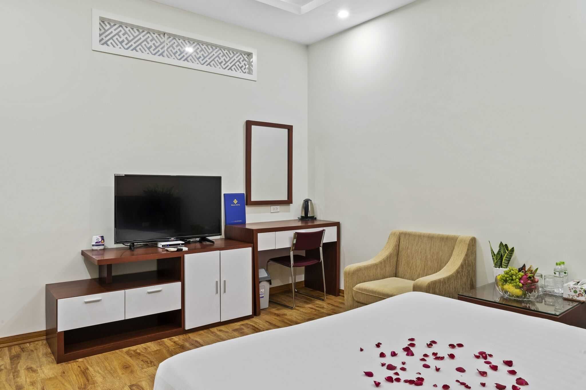 Quarto de hotel com cama, escrivaninha com televisão, cadeira e pequena mesa com planta e fruteira.