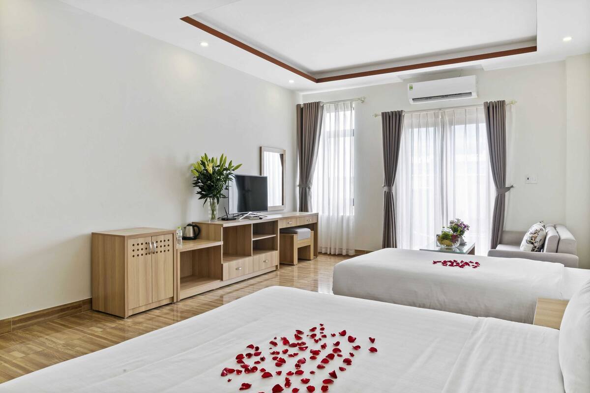 A hotel room with two beds, a desk with a TV, a plant, and a window with curtains.