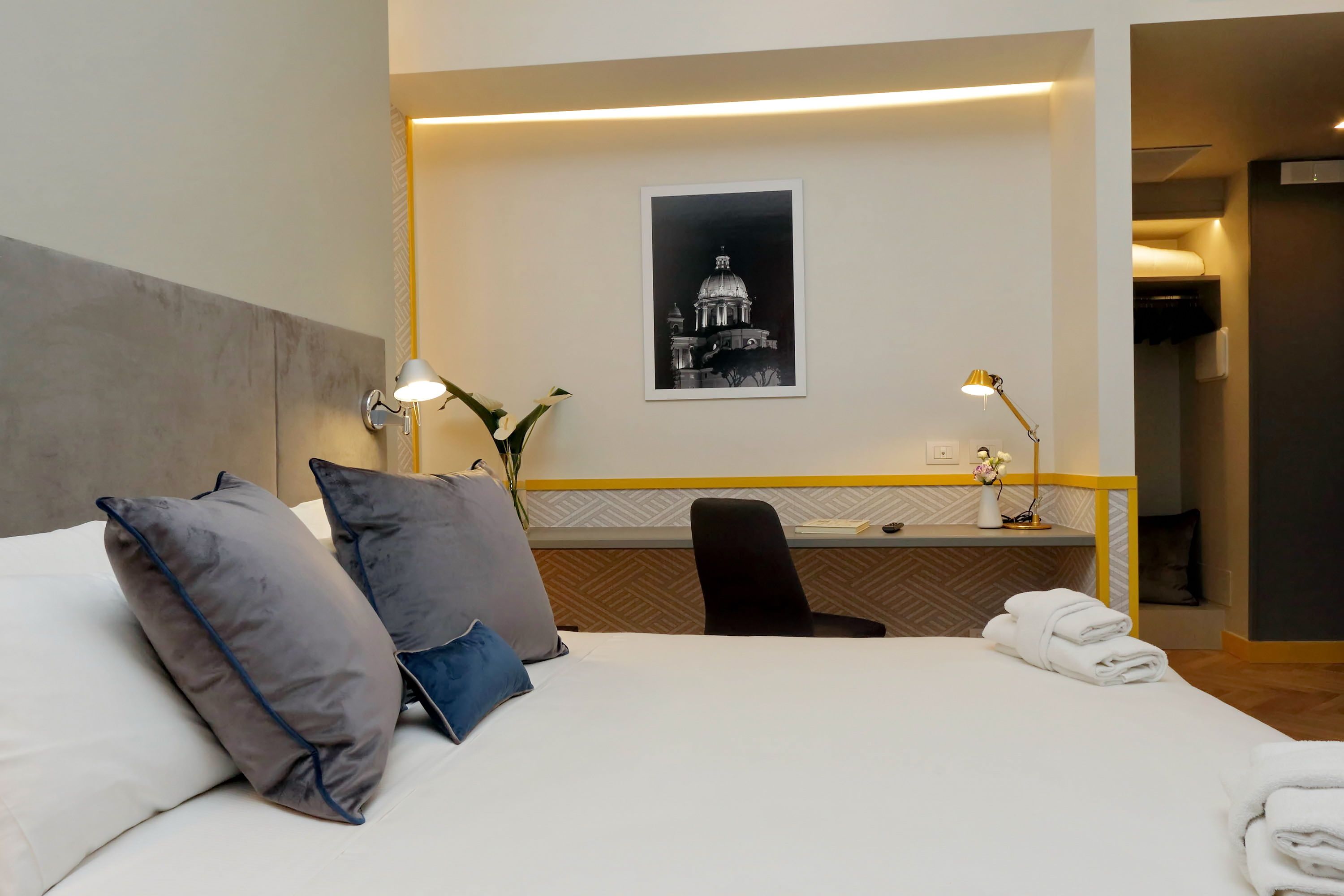 Ein Hotelzimmer mit Bett, Kissen, Schreibtisch, Stuhl, Lampe, einem gerahmten Bild an der Wand und einem Handtuch auf dem Bett.
