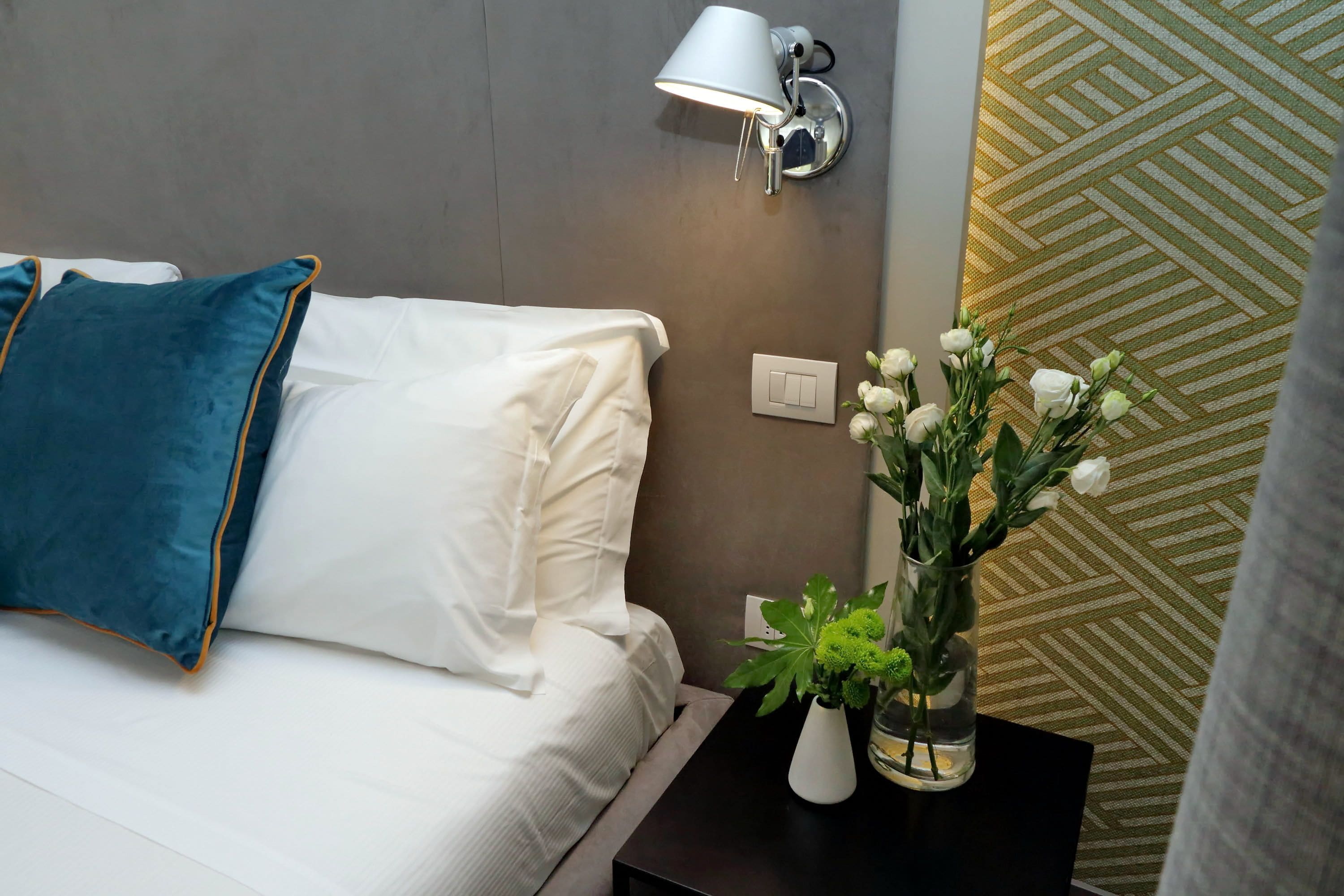 Una cama bien hecha con almohadas blancas y azules, una lámpara de noche, un florero con flores y una pared estampada.