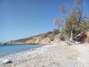 A pebbled beach with clear blue water, rocky cliffs, and sparse vegetation. - Kykladitiko Spiti (Syros)