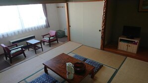 Une pièce de style japonais traditionnel, avec un sol recouvert de tatamis, une petite table sur laquelle se trouve une bouilloire, un téléviseur posé sur un meuble et une porte coulissante.