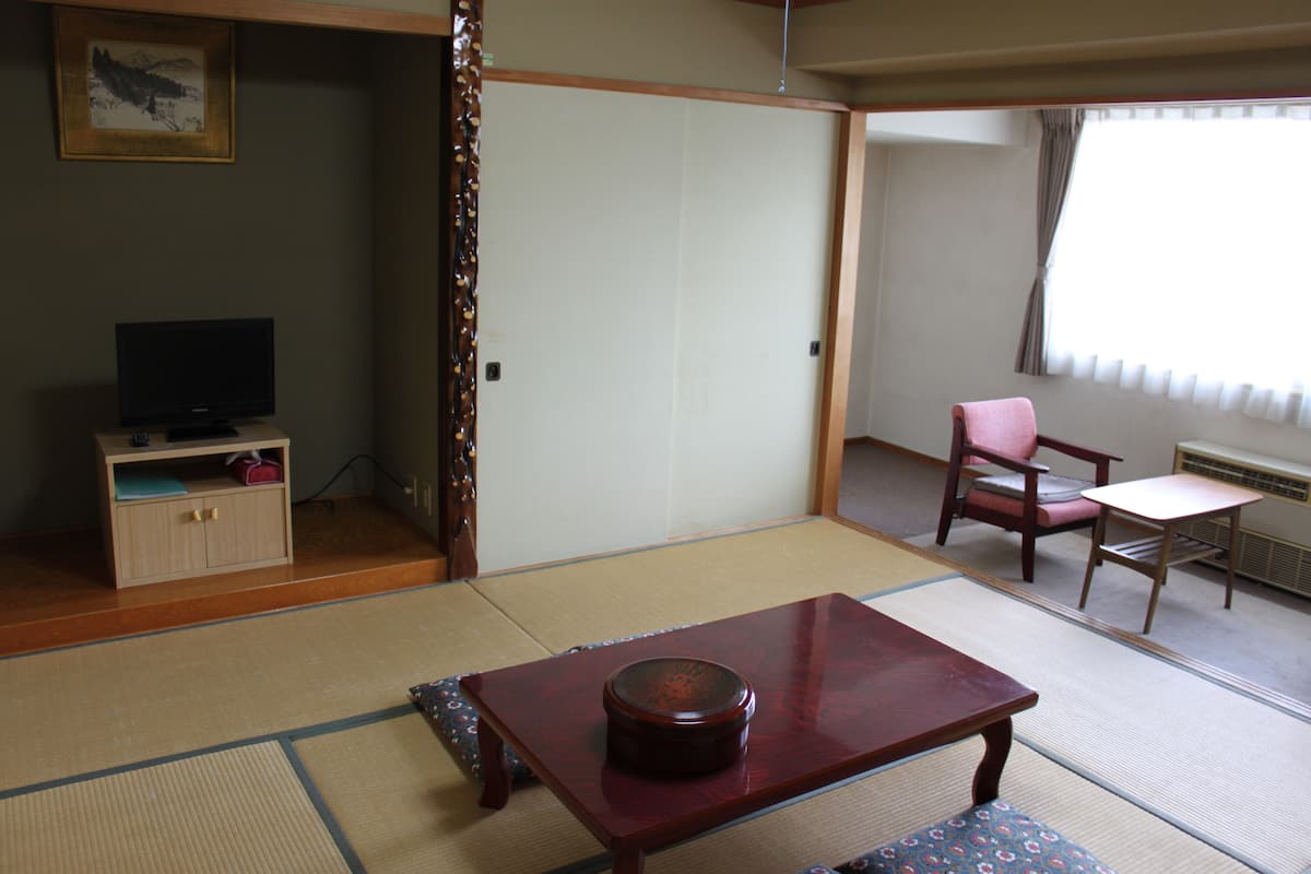 A traditional Japanese room with tatami flooring, a low table with a round object, a television on a cabinet, and a chair with a cushion.