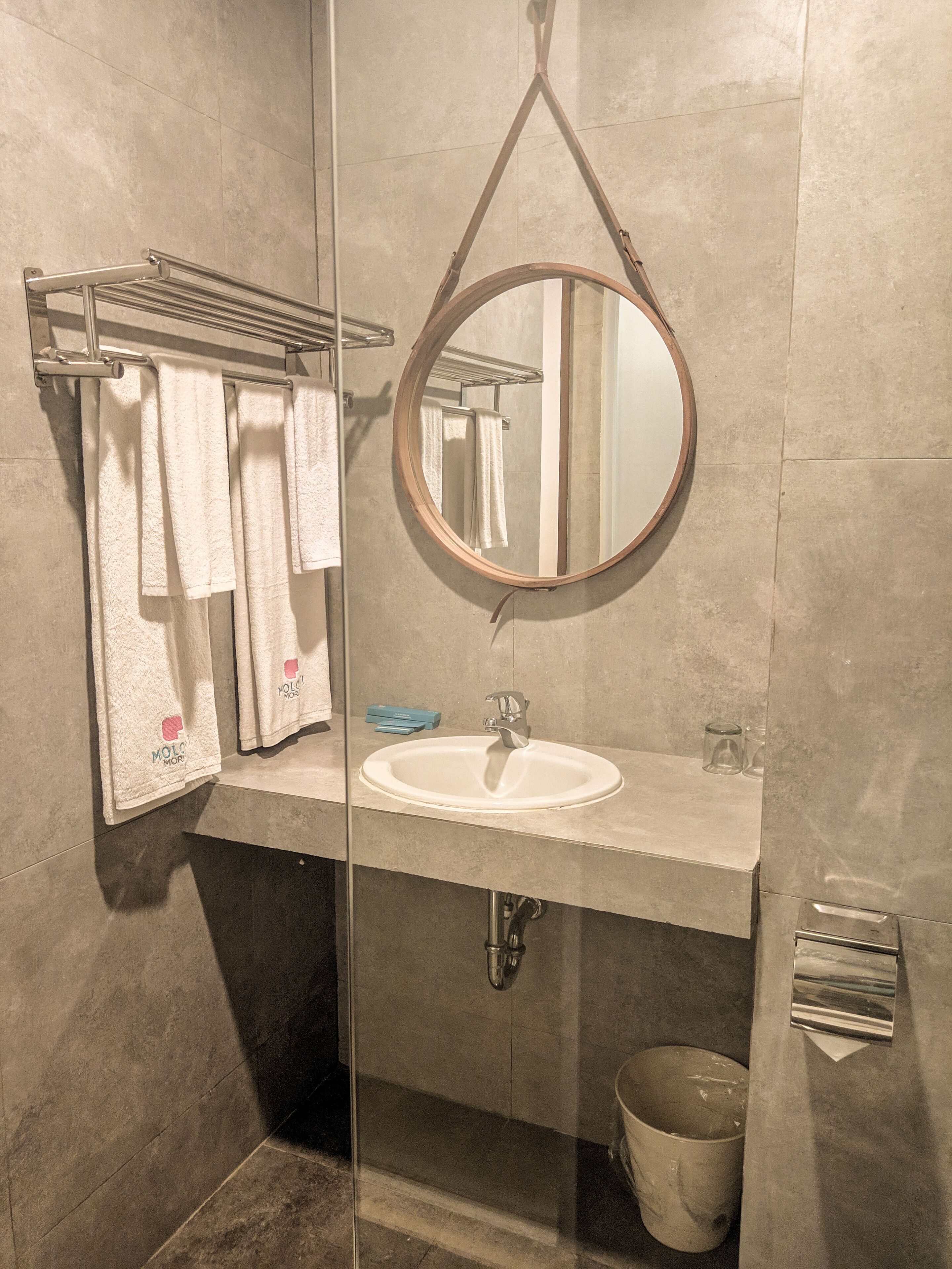 Une salle de bain moderne dotée d’une douche à parois de verre, d’un miroir rond, d’un lavabo blanc et d’un porte-serviettes sur lequel sont disposées plusieurs serviettes.