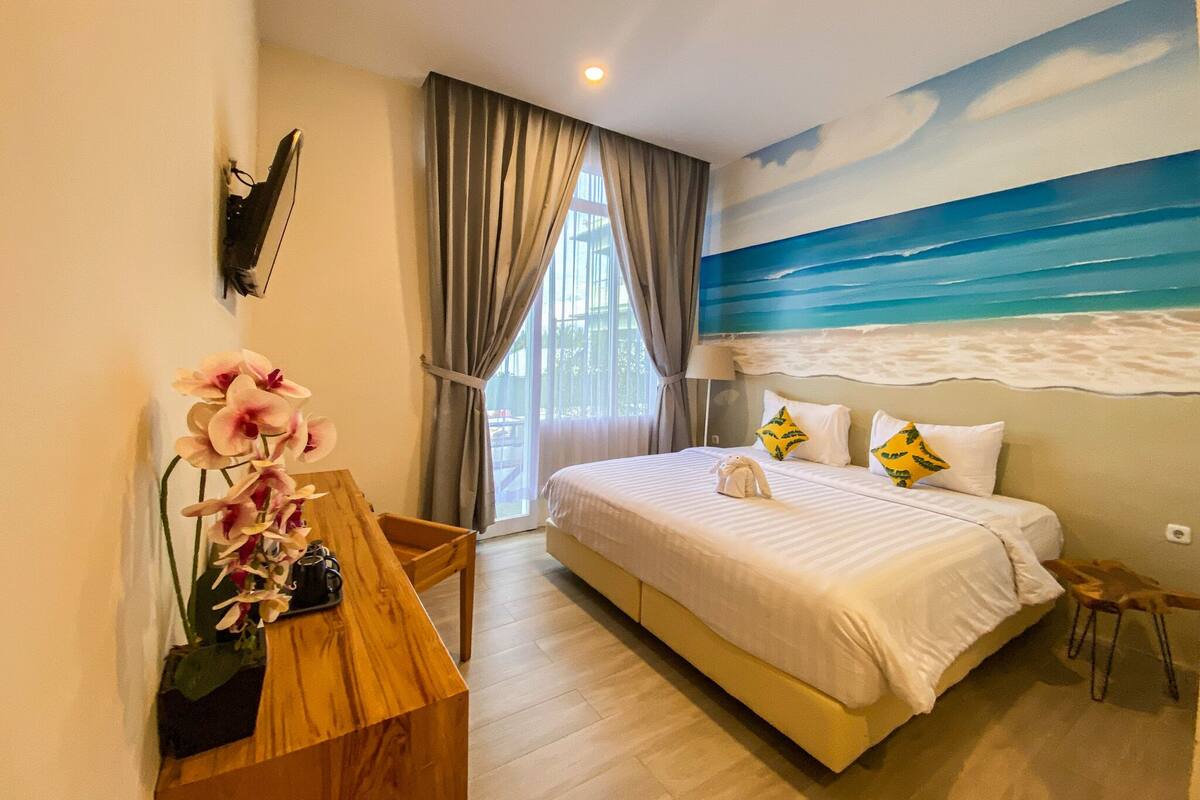 Ein Schlafzimmer mit einem Bett, einem Schreibtisch mit Blumen in einer Vase, einem Gemälde vom Strand und Blick ins Freie.