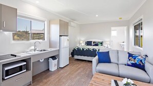 A compact room with a kitchenette, a bed with a blue and white quilt, and a grey sofa.