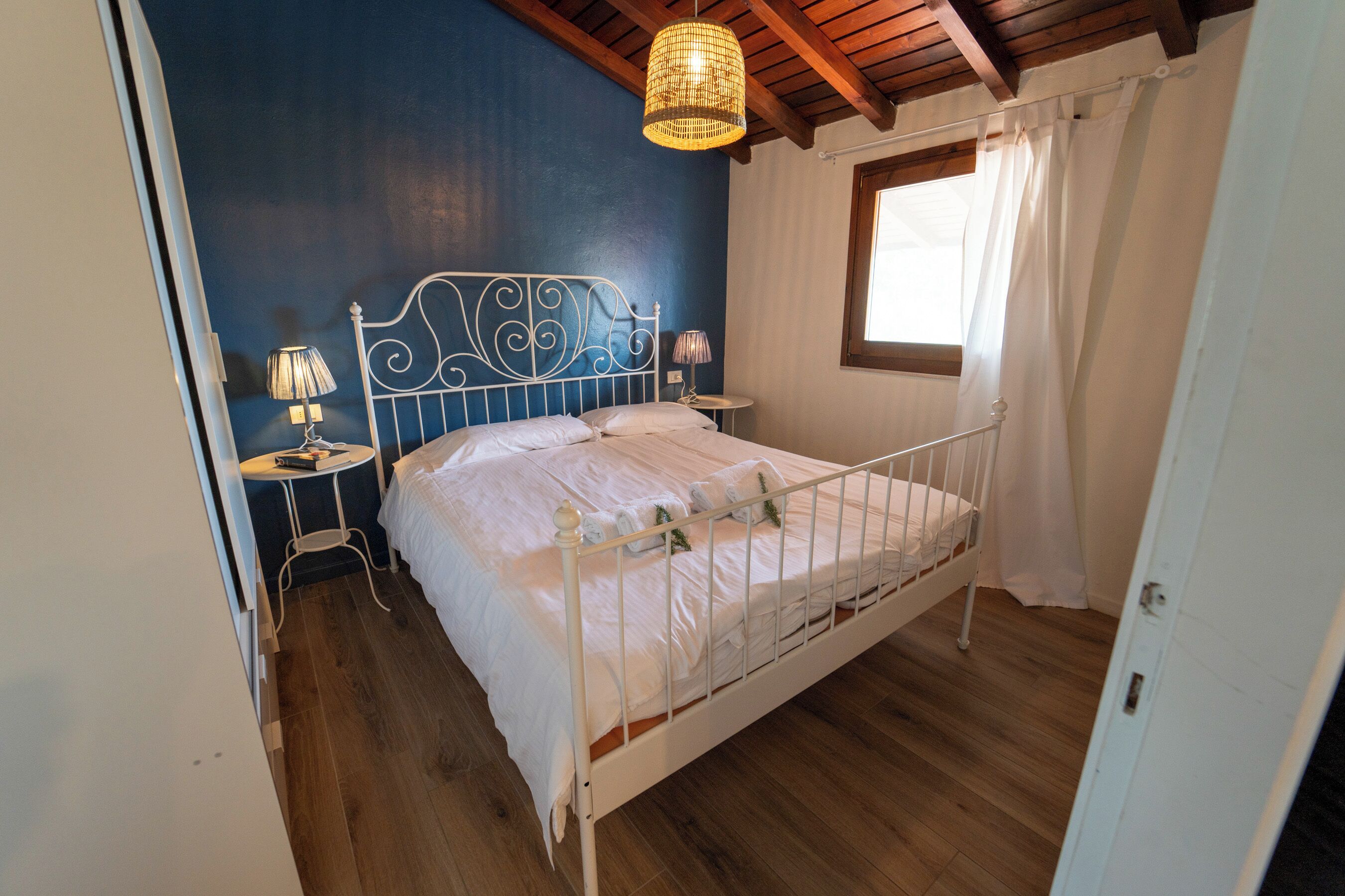 A bedroom with a metal bed frame, white bedding, a small bedside table with a lamp, and a window with curtains.