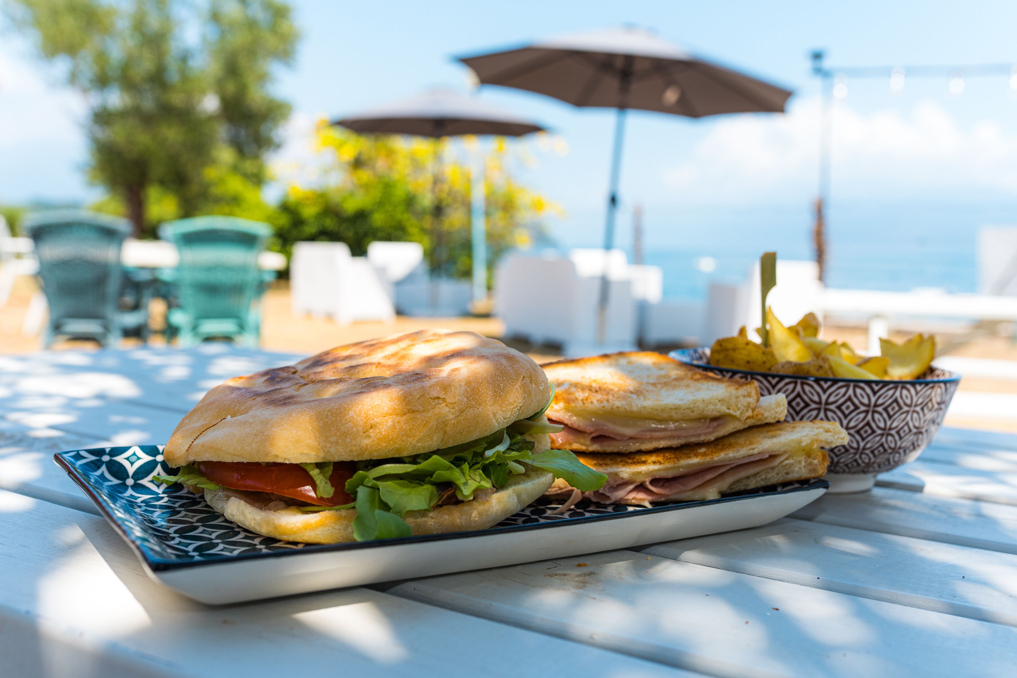 Een sandwich en frieten op een tafel, met uitzicht op zee en parasols op de achtergrond.