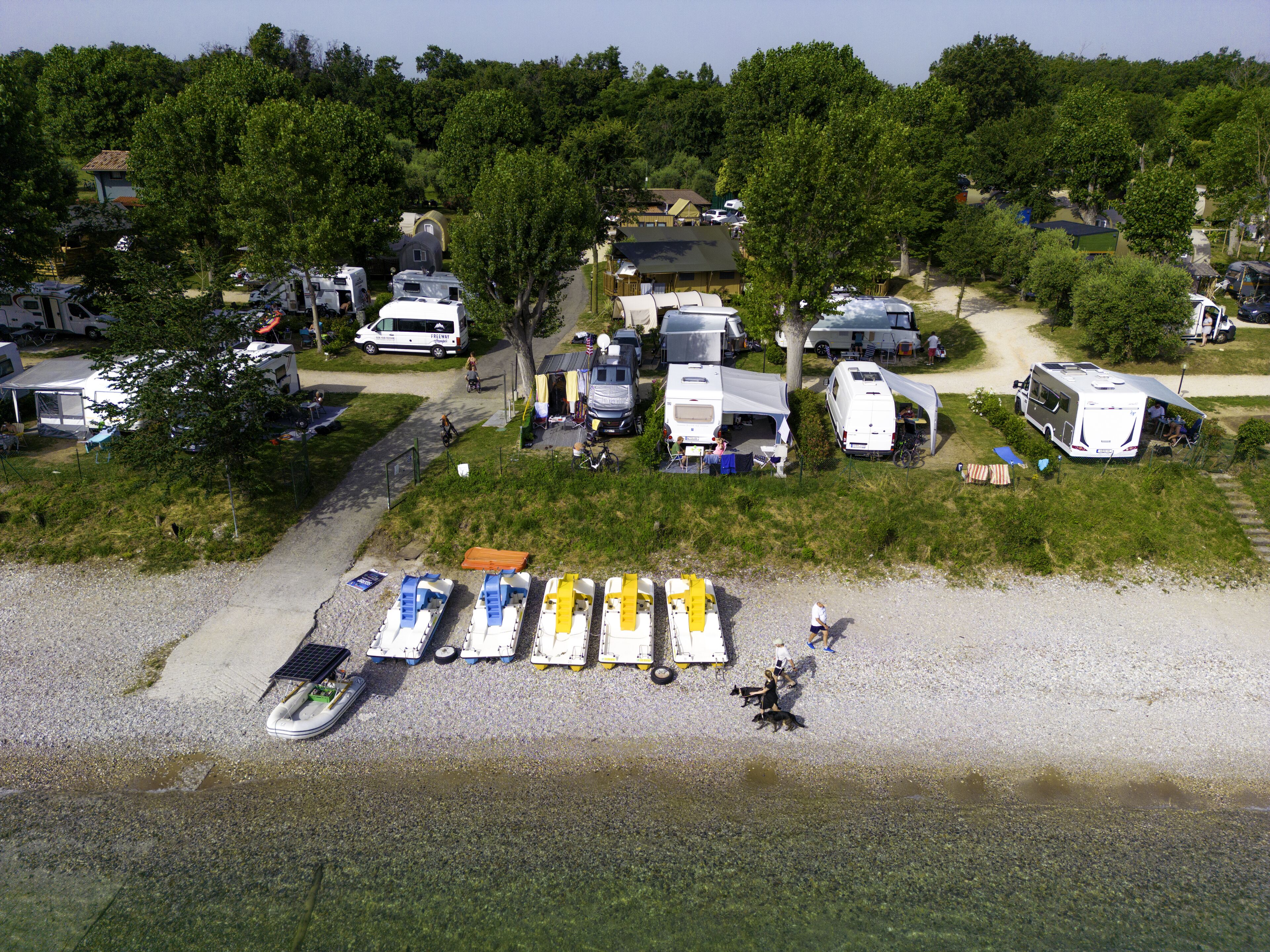 Un terrain de camping où sont stationnés des camping-cars et des remorques sur une zone herbeuse, près d’une étendue d’eau.