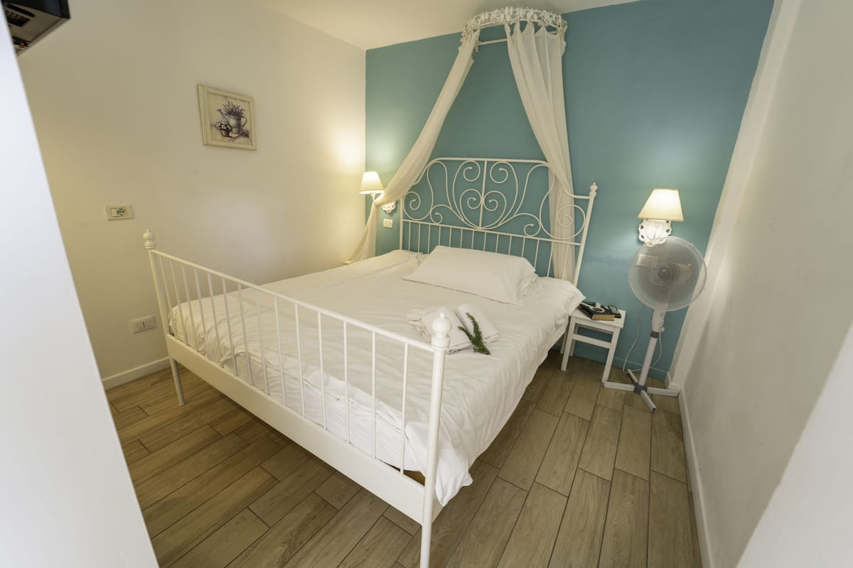 A white metal four-poster bed with a canopy, white bedding, a bedside table with a lamp, and a standing fan.