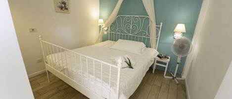 A white metal four-poster bed with a canopy, white bedding, a bedside table with a lamp, and a standing fan.