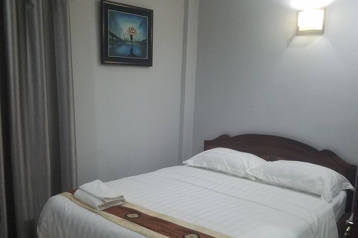 Una cama bien hecha con una colcha blanca y marrón, un cuadro en la pared y una ventana con una lámpara.
