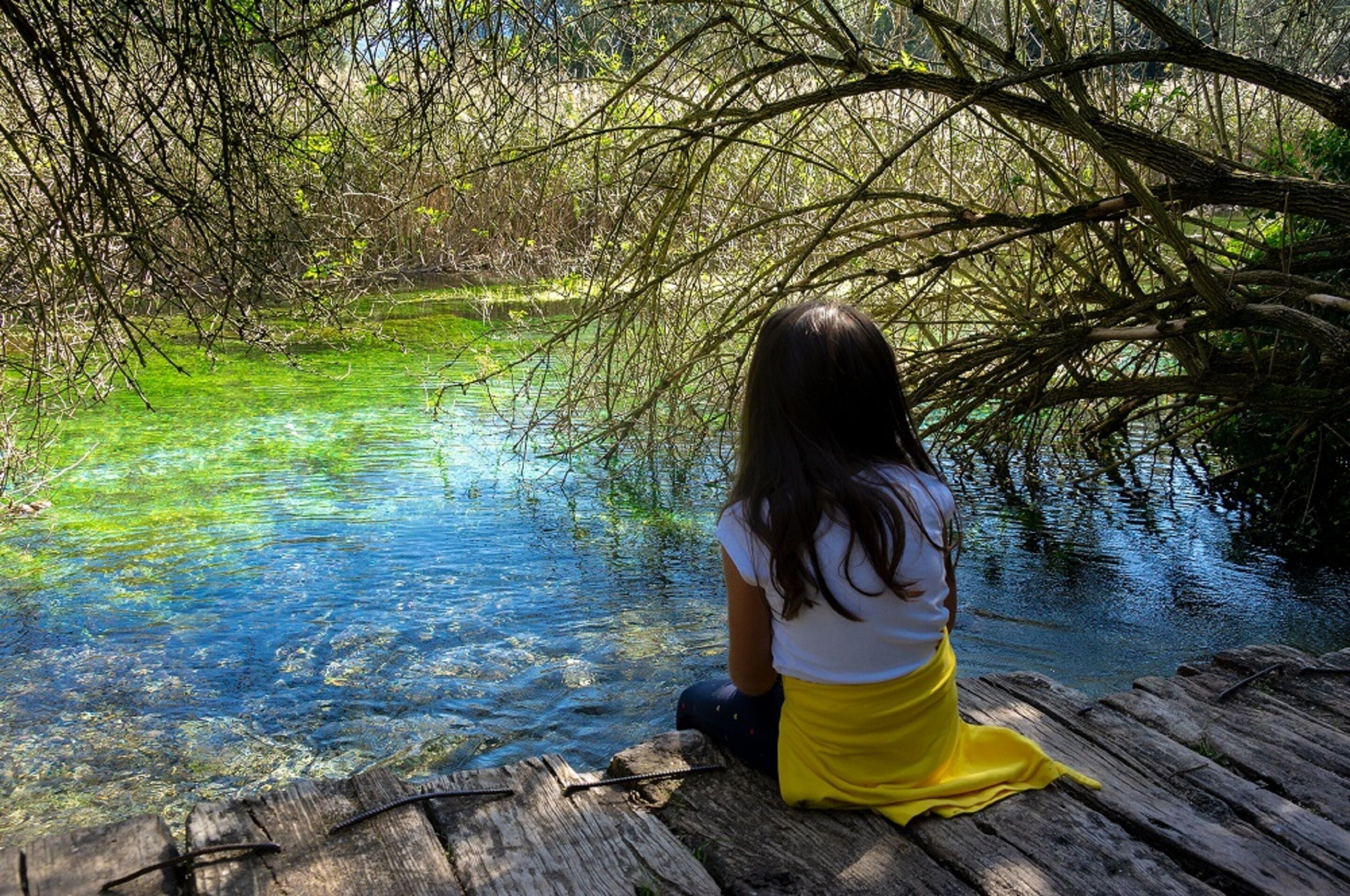 A person sitting on a wooden dock by a clear water body surrounded by dense foliage.