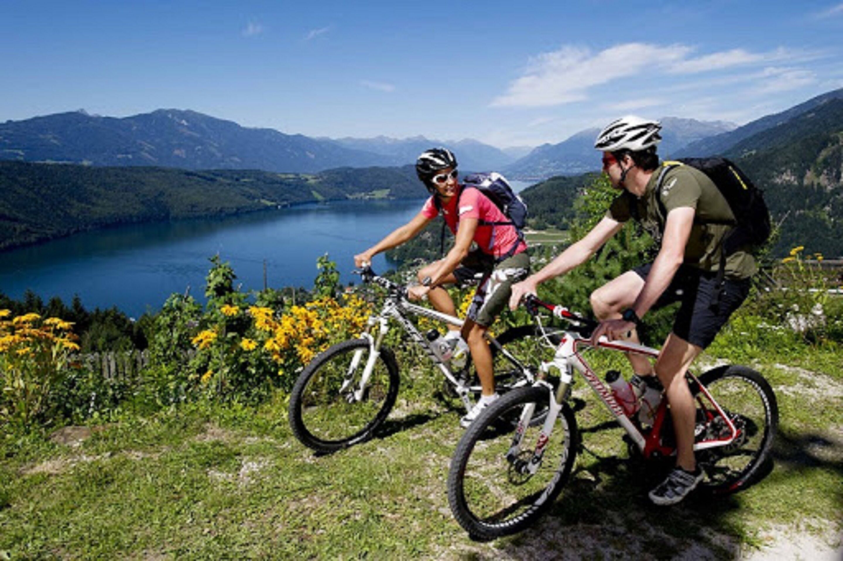 Dos ciclistas en un sendero de montaña con un lago y montañas al fondo.