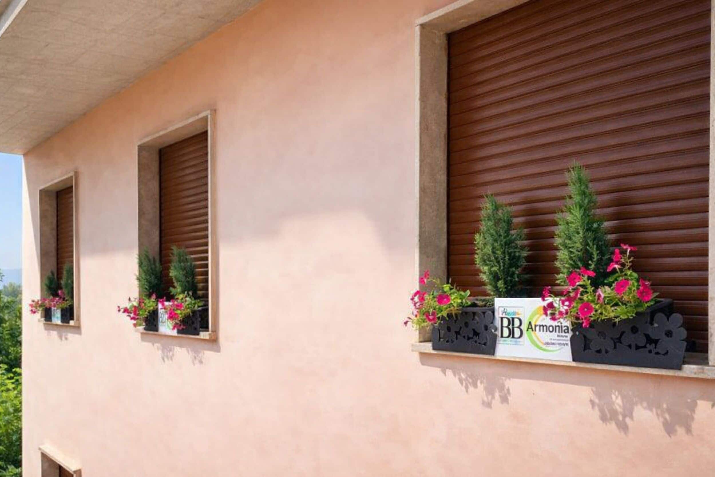 Un edificio con tres ventanas, cada una con un persiano. Hay macetas con flores rosadas y plantas verdes en los alféizares.
