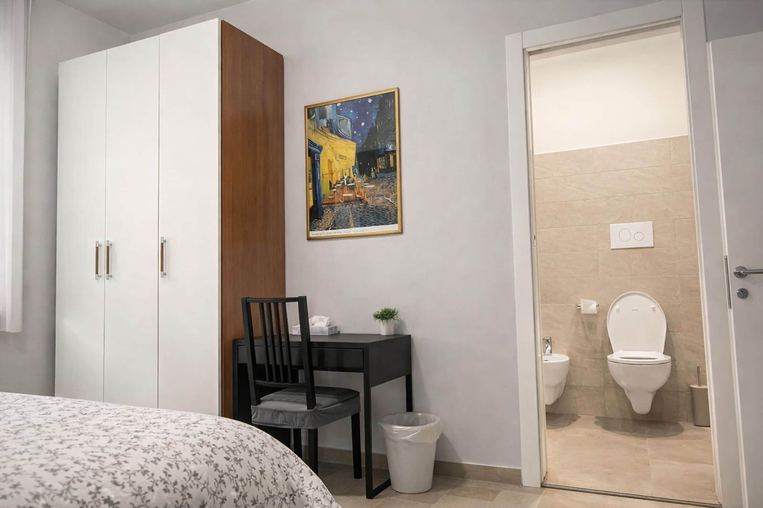A bedroom with a bed, a desk with a chair, a wardrobe, a painting on the wall, and a bathroom with a toilet and bidet.
