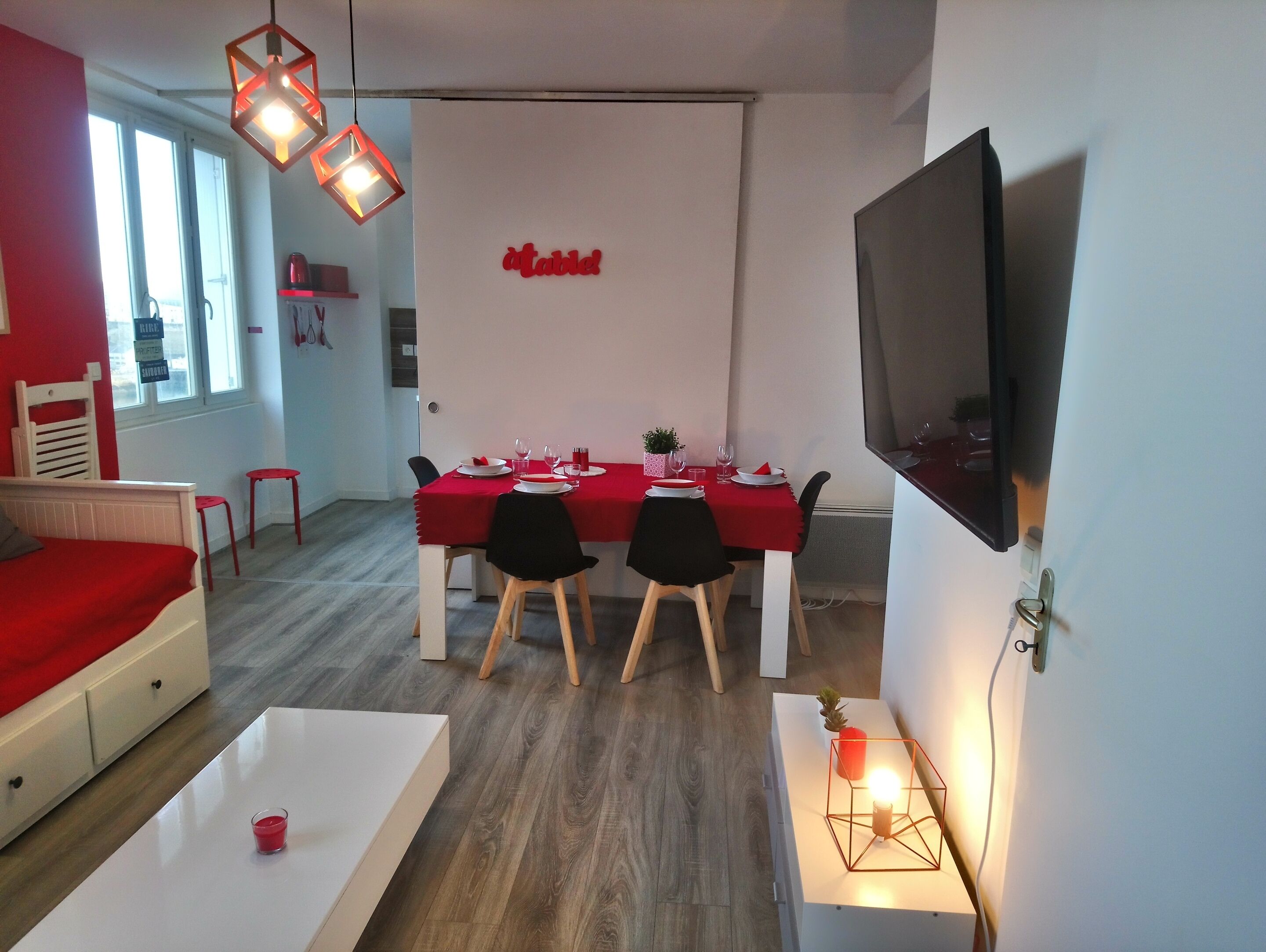 A modern dining area with a red tablecloth, two black chairs, and a white table. A flat-screen TV is mounted on the wall to the right.