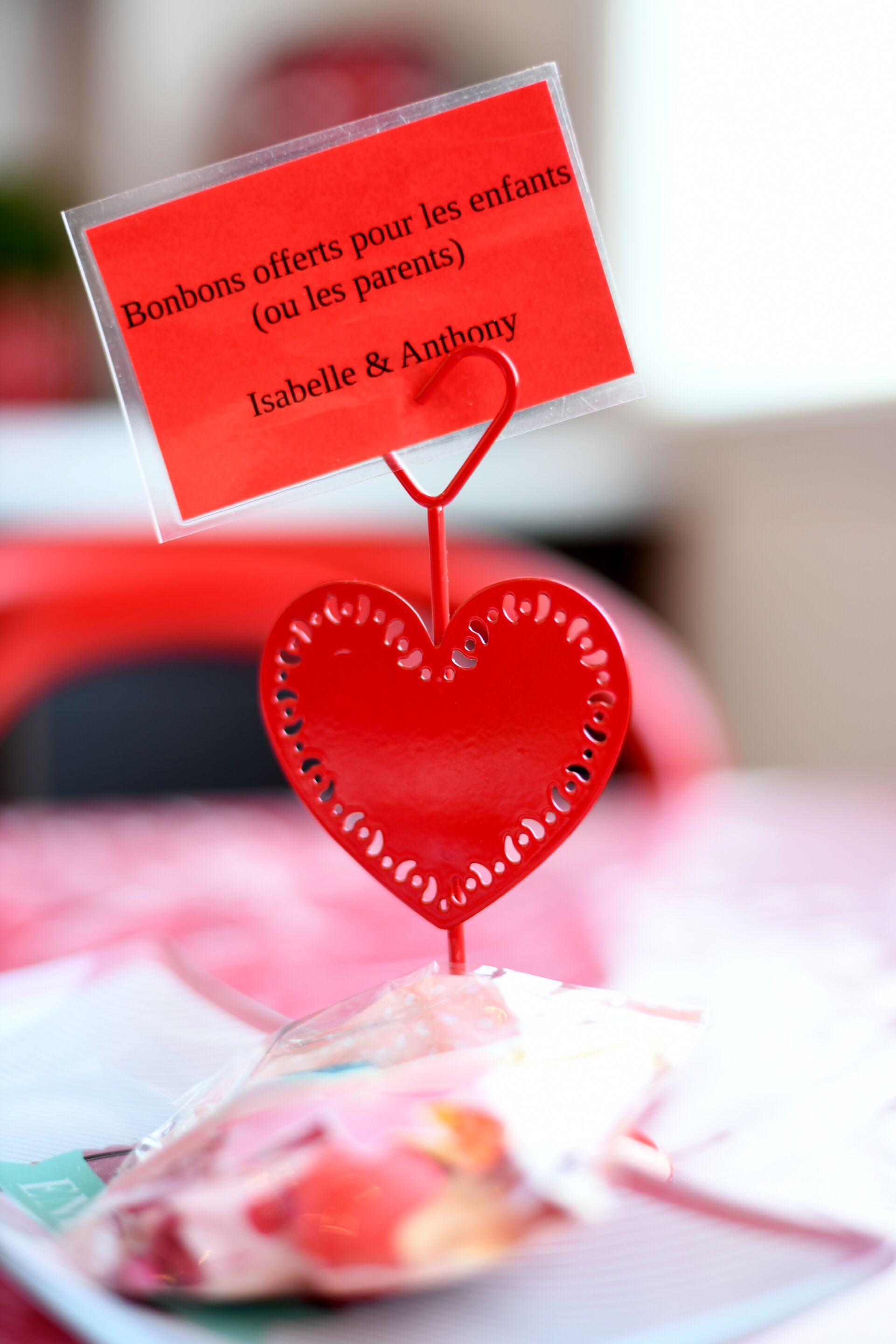 A red heart-shaped ornament with a sign attached, placed on a table with a blurred background.