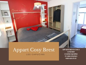 A bedroom with a red accent wall, a bed with a heart-shaped pillow, a wardrobe, and a small table with a lamp. - Appart Cosy Brest Les Capucins (Brest)