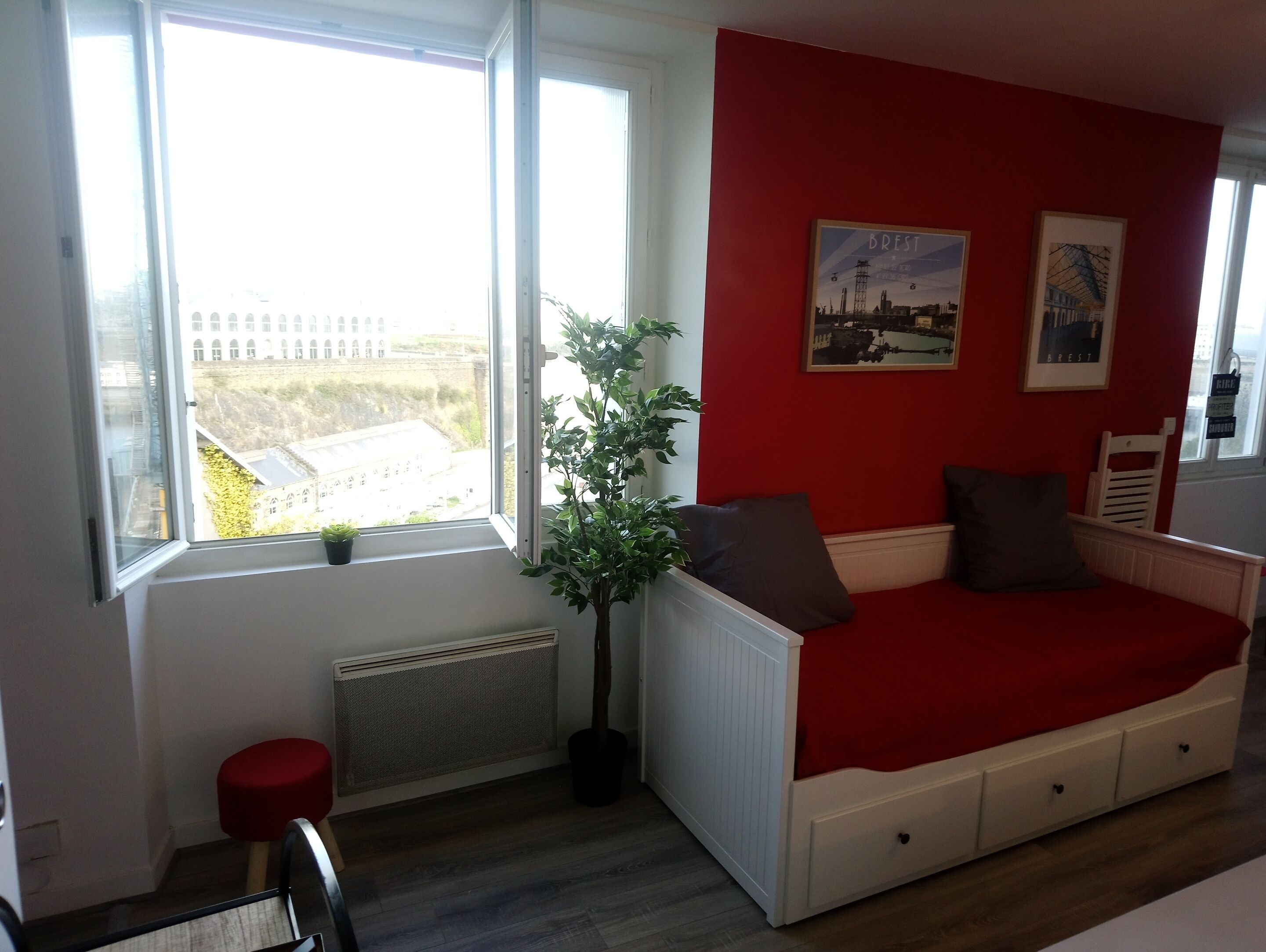 Una habitación pequeña y luminosa con una pared roja, una ventana con vista a edificios, un radiador, una planta y una cama con cubierta roja.